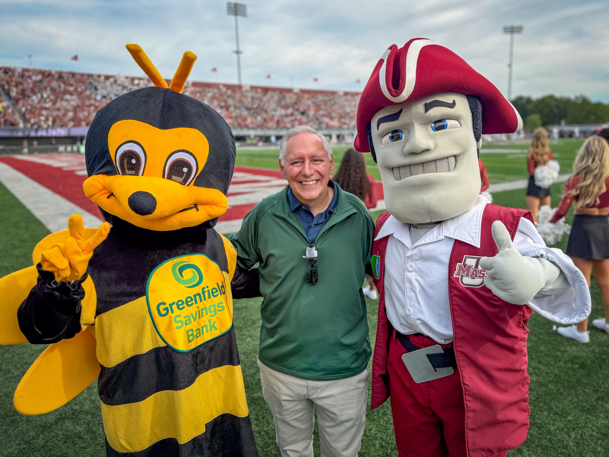 Peter Albero standing in between the GSB bee mascot and the UMass Minuteman mascot on the football field at McGuirk Stadium.