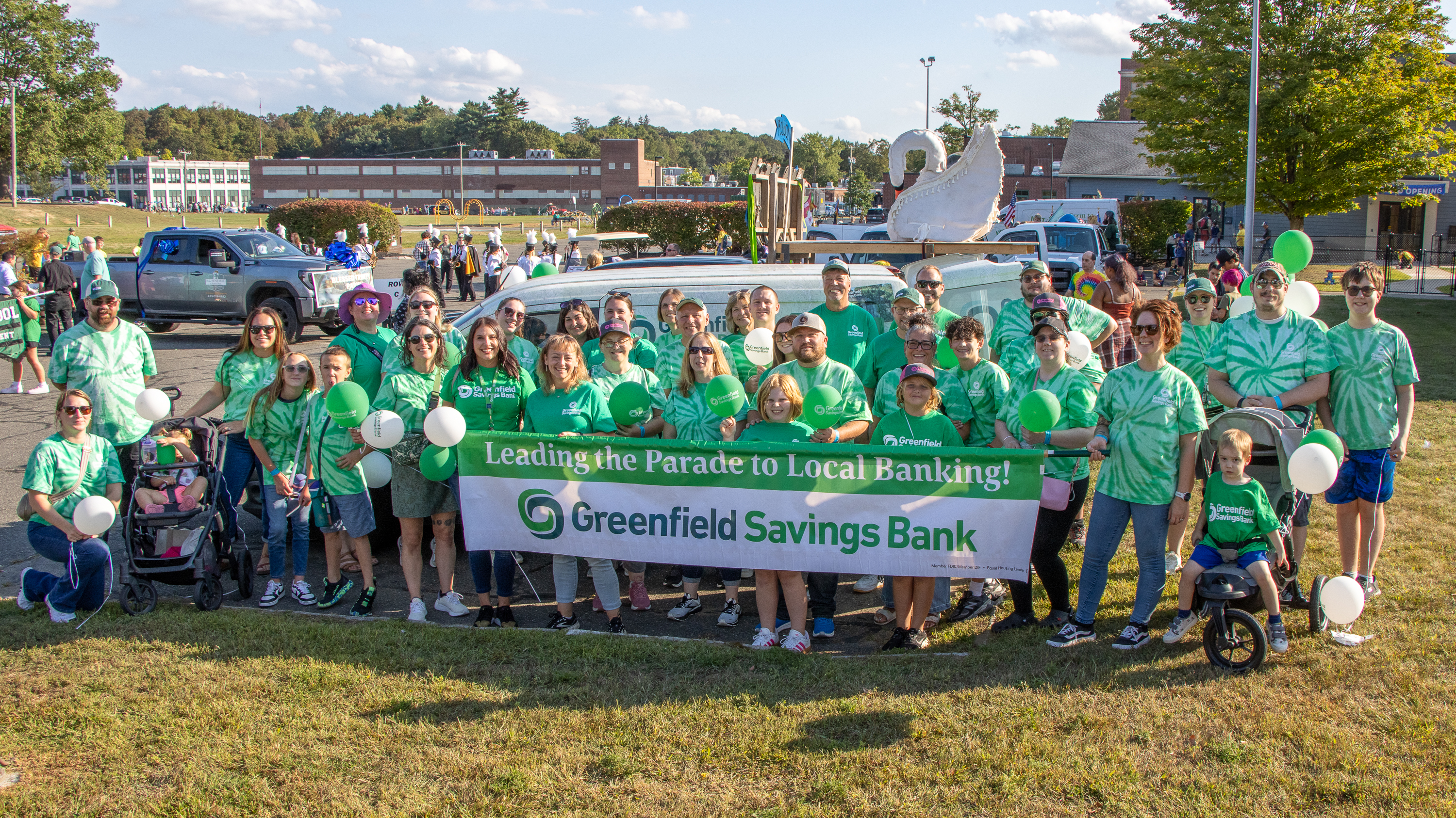A large group of Greenfield Savings Bank employees, family members, and community participants gather outdoors for a parade, wearing matching green tie-dye shirts and holding green and white balloons. The group stands in front of a parade float featuring a large white swan sculpture and a banner reading, “Leading the Parade to Local Banking! Greenfield Savings Bank.” Parade vehicles, spectators, and buildings are visible in the background on a sunny day.