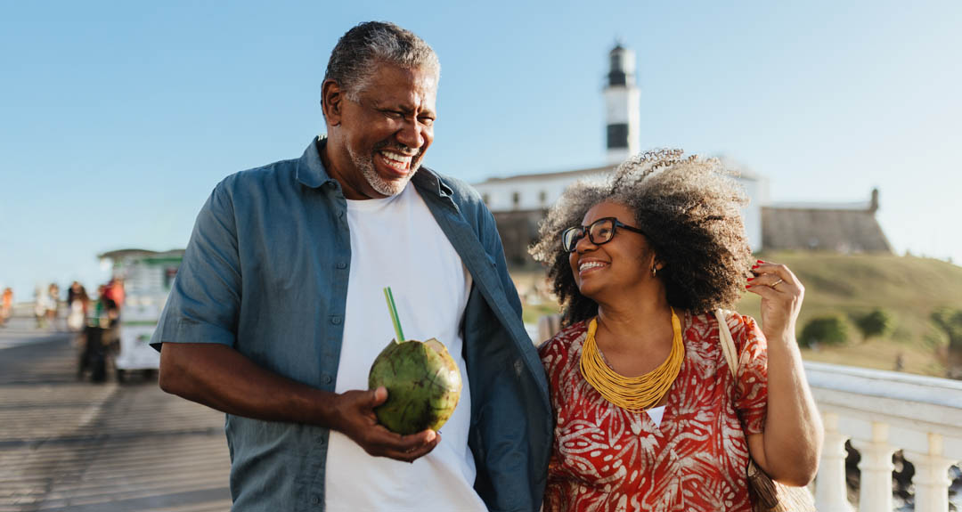 A middle aged couple is on vacation. There is a lighthouse in the background and the man is carrying a coconut with a straw in it.