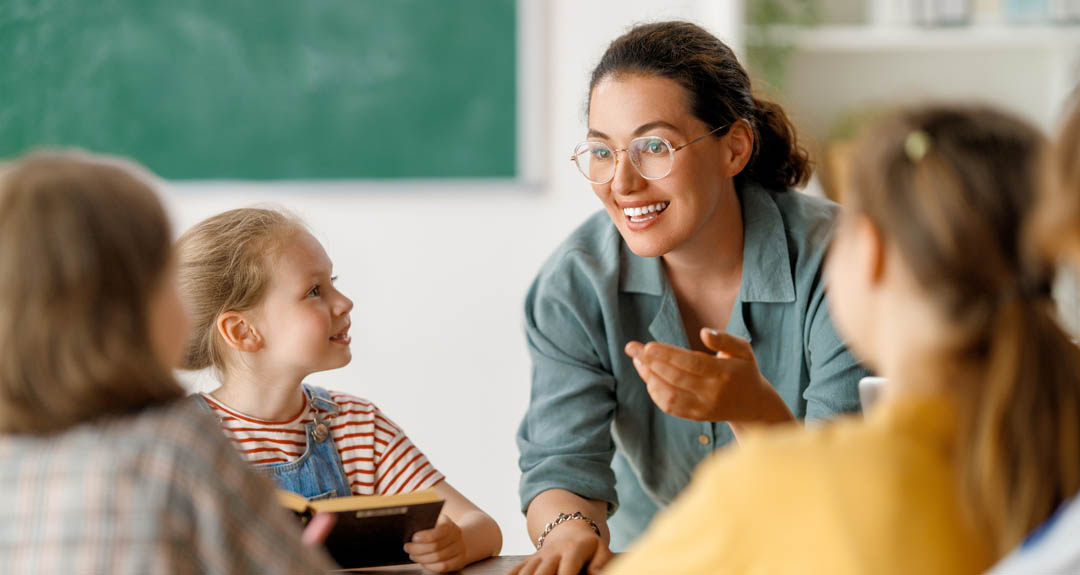 A brown haired woman who appears to be a teacher is in a green button down, and is teaching several young students. 