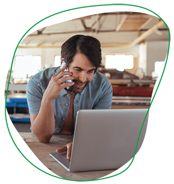 This image is a photo of a man on the phone, looking at his laptop, with a factory in the background. The man has brown hair and wears a denim short-sleeved shirt. The image is within a freeform element.