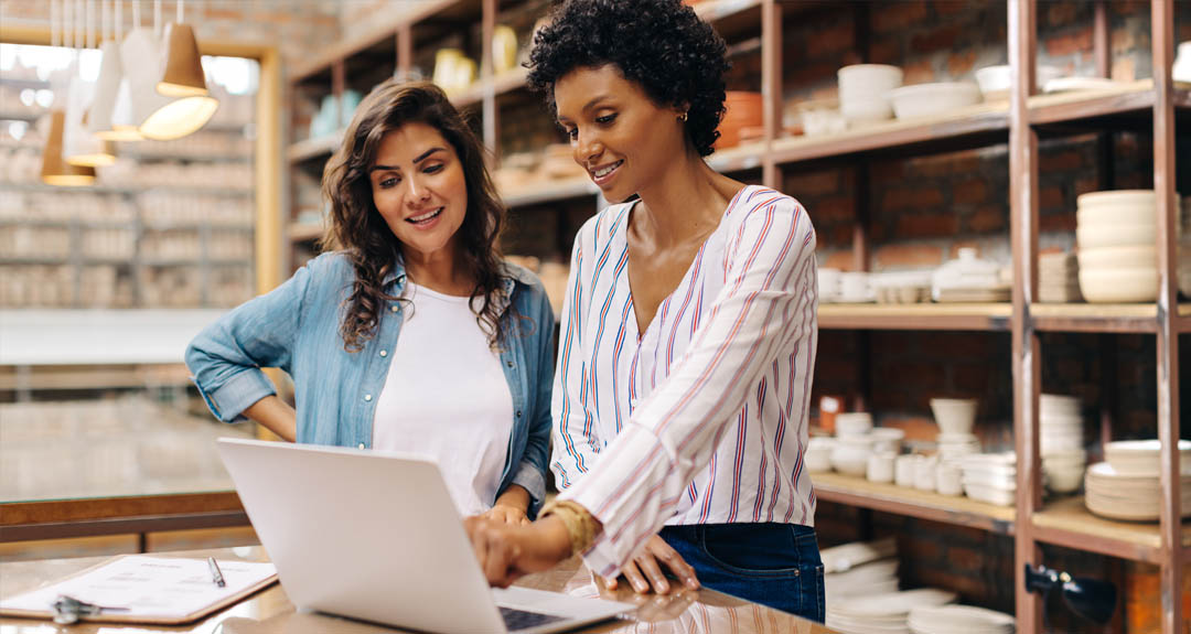 This image reflects two women working in a small business. The woman on the left has fair skin and long brown hair. She is wearing a blue blouse with a white t-shirt. The woman on the right has darker skin and short, curly, dark brown hair. She is wearing a white blouse with a v-neck. They are looking at a laptop and smiling. There is a shelf of pottery in the background.