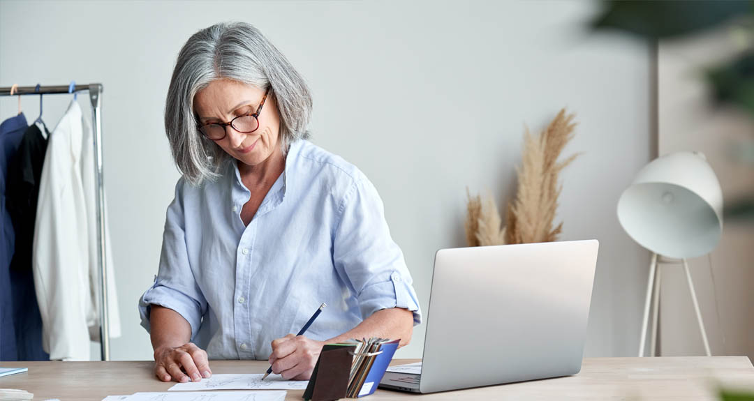 A gray haired business woman in a button-down blouse writes something in front of a laptop. 