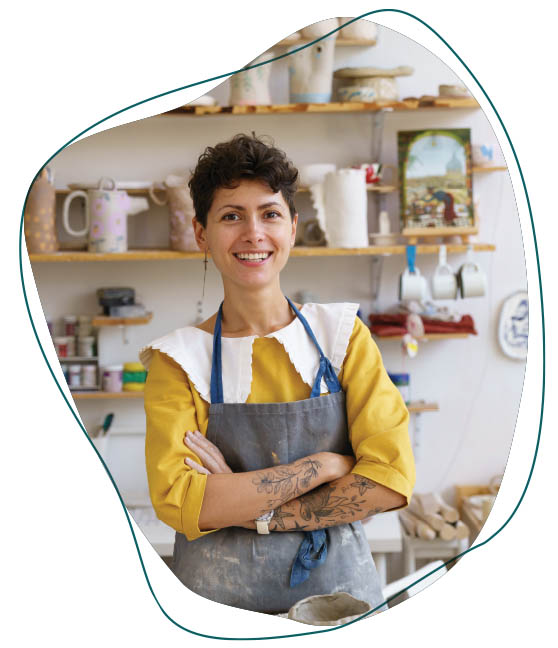 Photo of a woman who appears to be a potter, wearing an apron. She has a yellow blouse with a large white collar. Examples of pottery are visible in the background.