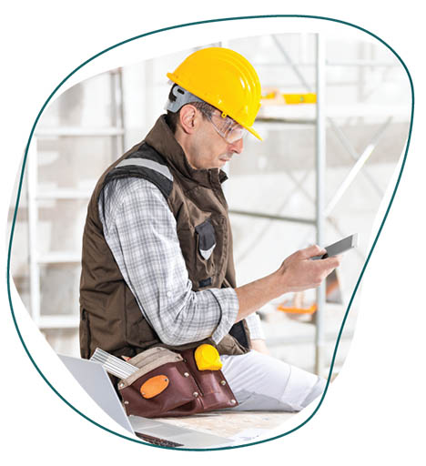 This is an image of a construction worker in a yellow hat. He is wearing a brown vest and has a grey plain flannel shirt under it. He has a brown work belt and is looking at his phone. 