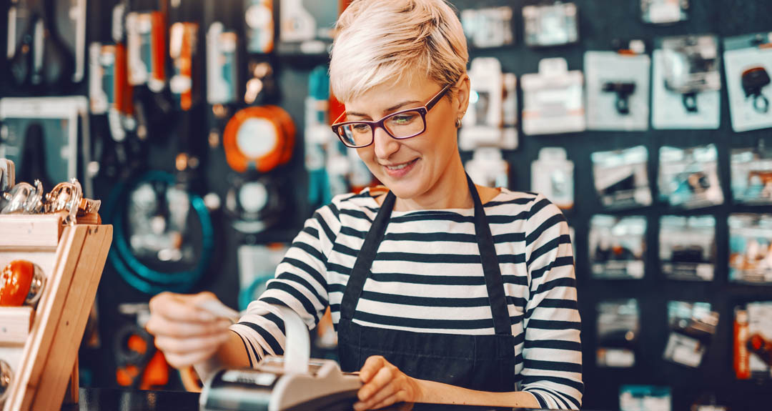 This is an image of a blonde woman with brown glasses who is a cashier at a business, looking at a receipt. She is wearing a black and white striped shirt and a black apron. 