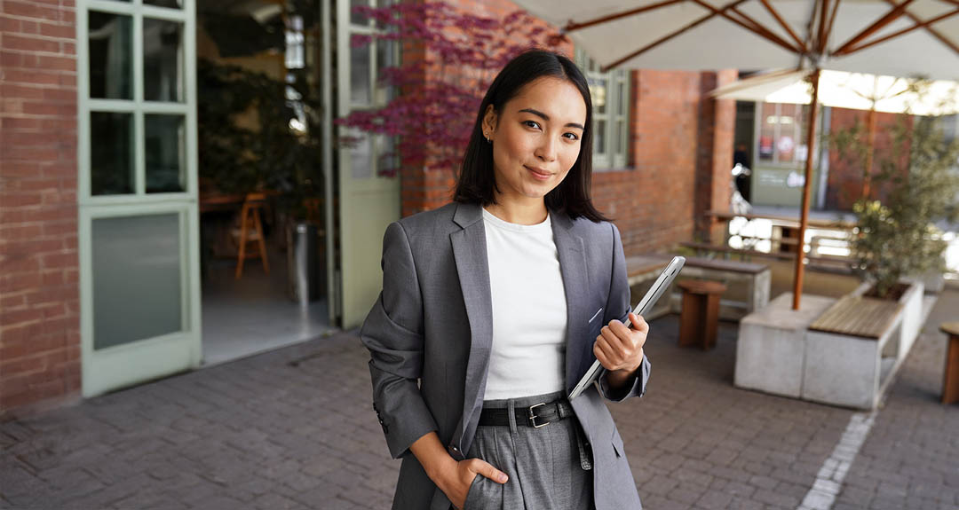 This image is a woman in front of a business, holding a laptop. She is wearing a grey suit with a white shirt. She has long, dark, straight hair.
