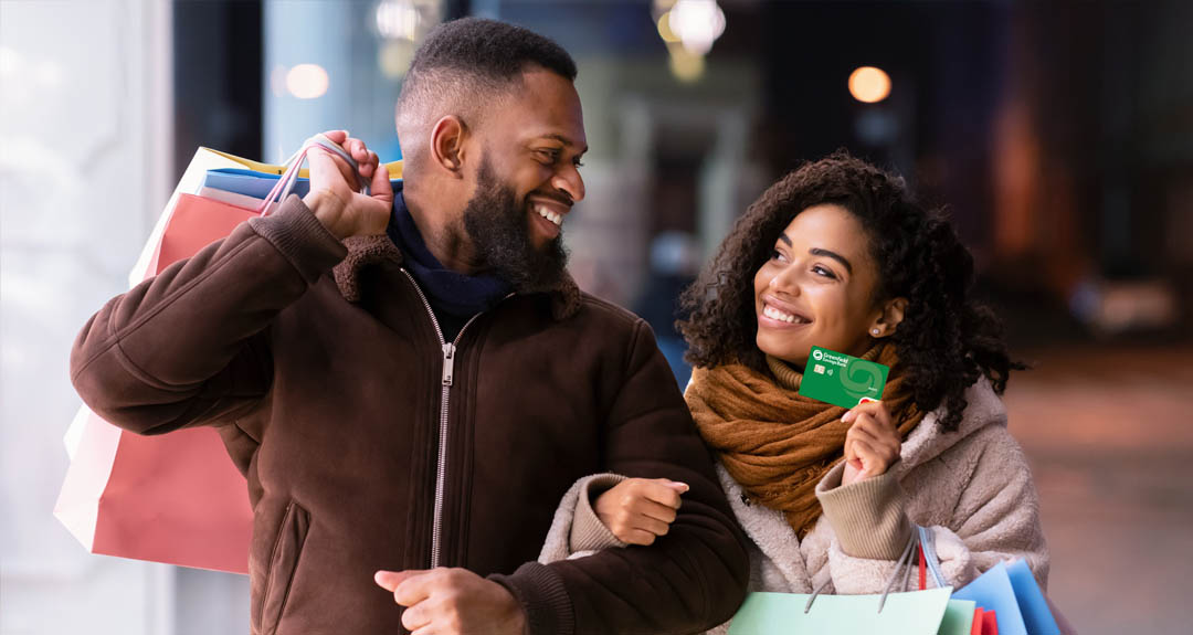 A young 20-something couple are walking down a trendy street at night, carrying shopping bags. The woman is smiling and holding a GSB debit card. 